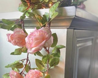 May include: A decorative garland featuring soft pink fabric roses and green leaves intertwined with small, warm-toned string lights. The floral vine hangs down, showcasing layered petals and realistic foliage attached to thin, flexible brown stems.