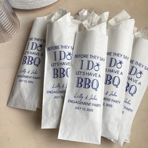 May include: White paper cutlery sleeves containing clear plastic forks, each printed with blue text reading BEFORE THEY SAY I DO LET'S HAVE A BBQ, Lilly & John ENGAGEMENT PARTY JULY 12, 2025, featuring a diamond ring icon above the word DO.