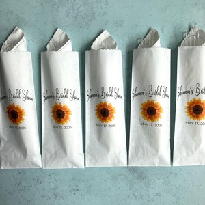 May include: Five white paper bags featuring a printed sunflower graphic, the text "Shannon's Bridal Shower", and the date "May 17, 2025". Each bag is folded at the top and contains a clear plastic utensil set visible at the opening of the paper sleeve.
