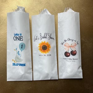 Op de afbeelding: Drie witte papieren zakjes met aangepaste ontwerpen. Het eerste zakje toont een gans, een blauwe ballon en de tekst Luka is ONE SILLY GOOSE. Het tweede toont een zonnebloem en Kat's Bridal Shower MAY 24, 2026. Het derde toont kersen en She's the Cherry on top Aniya's Baby Shower May 16, 2026.
