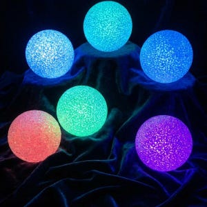 May include: Five spherical lamps with a textured, crackled surface finish. Each globe emits a distinct vibrant glow, including shades of cyan, blue, green, pink, and purple, creating a luminous display of light against a dark, draped fabric.