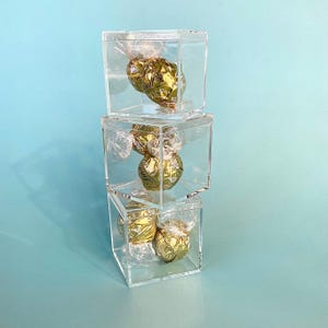 May include: Three clear acrylic boxes stacked on top of each other, each containing individually wrapped gold foil candies. The boxes are square and transparent, with the candies visible inside. The background is a solid light blue.