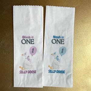 It displays two white rectangular items featuring a goose illustration, the text Olivia is ONE or Noah is ONE, a balloon with the number 1, and the words SILLY GOOSE. One Silly Goose Birthday Girl Personalize Silverware Bag Boy Baby 1st Birthday Favor Custom Cutlery Bag Table Decor Paper Napkin Holder Idea
