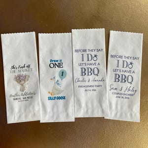 Op de afbeelding: Vier witte papieren zakjes met geschulpte bovenranden. Het eerste zakje heeft een bloemenboeket en de tekst She's Fresh off THE MARKET Heather AshlieAnn's BRIDAL SHOWER June 27, 2026. Het tweede toont een gans en ballon met de tekst Drew is ONE SILLY GOOSE. Het derde en vierde zakje tonen BEFORE THEY SAY I DO LET'S HAVE A BBQ met de namen Charlie & Amanda of Sam & Haley, evenementtypes en datums.