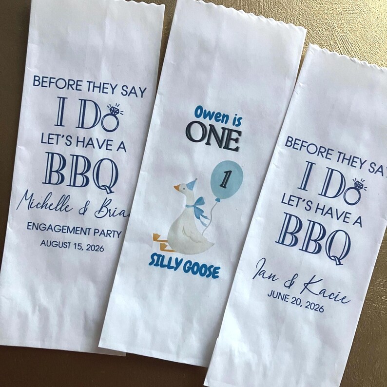Op de afbeelding: Drie witte papieren zakken met gekartelde bovenranden. Twee zakken hebben blauwe tekst met BEFORE THEY SAY I DO LET'S HAVE A BBQ, namen zoals Michelle & Brian of Jan & Kacie en datums. E&eacute;n zak toont een gans en de tekst Owen is ONE.