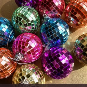Op de afbeelding: Een verzameling kleine, bolvormige discobalornamenten bedekt met reflecterende, veelkleurige vierkante spiegeltjes in tinten paars, roze, blauw, groen, oranje en goud, elk voorzien van een zilveren ophanglus aan de bovenzijde.