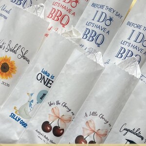 Op de afbeelding: Een verzameling witte papieren bestekzakjes met diverse bedrukte ontwerpen, waaronder teksten zoals Before They Say I Do Let's Have A BBQ, Luka is One en She's the Cherry on Top, naast afbeeldingen van zonnebloemen, ganzen en kersen.