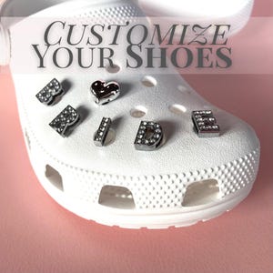 May include: A white foam clog shoe featuring silver-toned, rhinestone-encrusted letter charms spelling the word BRIDE and a silver heart charm inserted into the ventilation holes. The text CUSTOMIZE YOUR SHOES is overlaid at the top of the image.