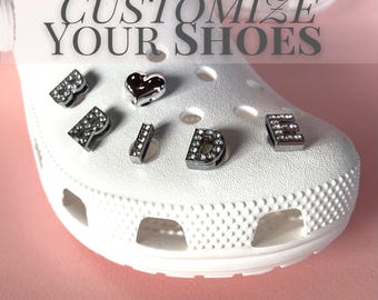 May include: A white foam clog shoe featuring silver-toned, rhinestone-encrusted letter charms spelling the word BRIDE and a silver heart charm inserted into the ventilation holes. The text CUSTOMIZE YOUR SHOES is overlaid at the top of the image.