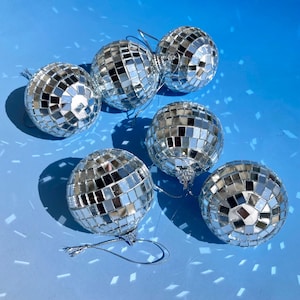 May include: A collection of five spherical ornaments covered in small, reflective silver mirror tiles. Each round ornament features a metallic hanging loop at the top, designed to catch and scatter light across the surrounding area.