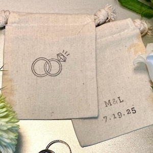May include: Two off-white cotton drawstring pouches. One pouch features a dark gray printed icon of two interlocking rings with a diamond. The other pouch displays the dark gray printed text M&L above the date 7.19.25 in a simple font.