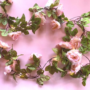 May include: A decorative string of artificial light pink roses featuring green fabric leaves and thin brown stems. The floral garland is interspersed with small integrated LED lights that emit a soft glow throughout the rose blossoms and foliage.