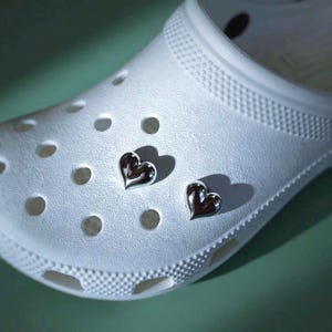 May include: A white foam clog shoe featuring two small, shiny, silver-toned heart-shaped charms inserted into the circular ventilation holes on the upper surface of the footwear. The charms have a smooth, reflective, metallic finish.