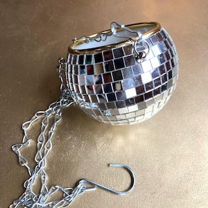 May include: A spherical hanging planter covered in small, reflective square mirror tiles. The planter features a gold-toned rim at the top opening and is suspended by a silver-colored metal chain that includes a hook for hanging purposes.