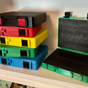 May include: A stack of five colorful plastic storage cases with black latches. The cases are black, red, yellow, green, and blue. The cases are stacked on a white shelf. A green plastic case with a black foam interior is open and sitting on the shelf.