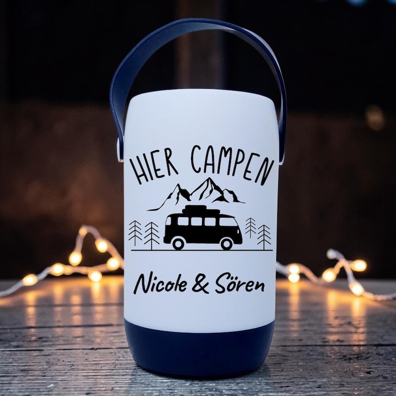 Peut inclure: Une lanterne de camping blanche avec une poign&eacute;e et une base bleu fonc&eacute;. La lanterne pr&eacute;sente du texte et des graphiques noirs, dont les mots "HIER CAMPEN", des montagnes, une camionnette et les noms "Nicole & S&ouml;ren".