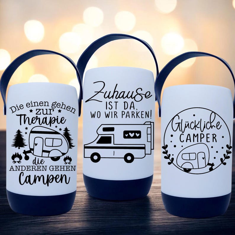 Peut inclure: Trois lanternes de camping blanches avec des poign&eacute;es et des bases noires. Chaque lanterne pr&eacute;sente un motif graphique et textuel noir diff&eacute;rent. L'une dit "Die einen gehen zur Therapie, die anderen gehen Campen". Une autre dit "Zuhause ist da, wo wir parken!" La troisi&egrave;me dit "Gl&uuml;ckliche Camper".