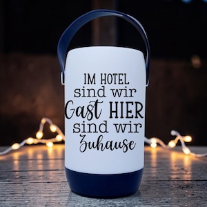 Peut inclure: Une lanterne blanche avec une base et une poign&eacute;e bleu marine. La lanterne porte l'inscription "IM HOTEL sind wir Gast HIER sind wir Zuhause" en noir. La lanterne est pos&eacute;e sur une surface en bois avec des guirlandes lumineuses en arri&egrave;re-plan.