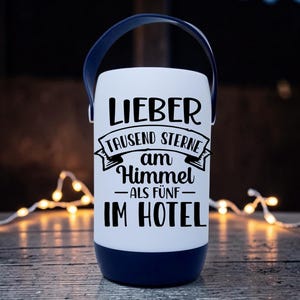 Peut inclure: Une lanterne blanche avec une poign&eacute;e et une base bleu marine. La lanterne porte un texte noir qui se lit "LIEBER TAUSEND STERNE AM HIMMEL ALS F&Uuml;NF IM HOTEL". La lanterne est pos&eacute;e sur une surface en bois avec des guirlandes lumineuses en arri&egrave;re-plan.
