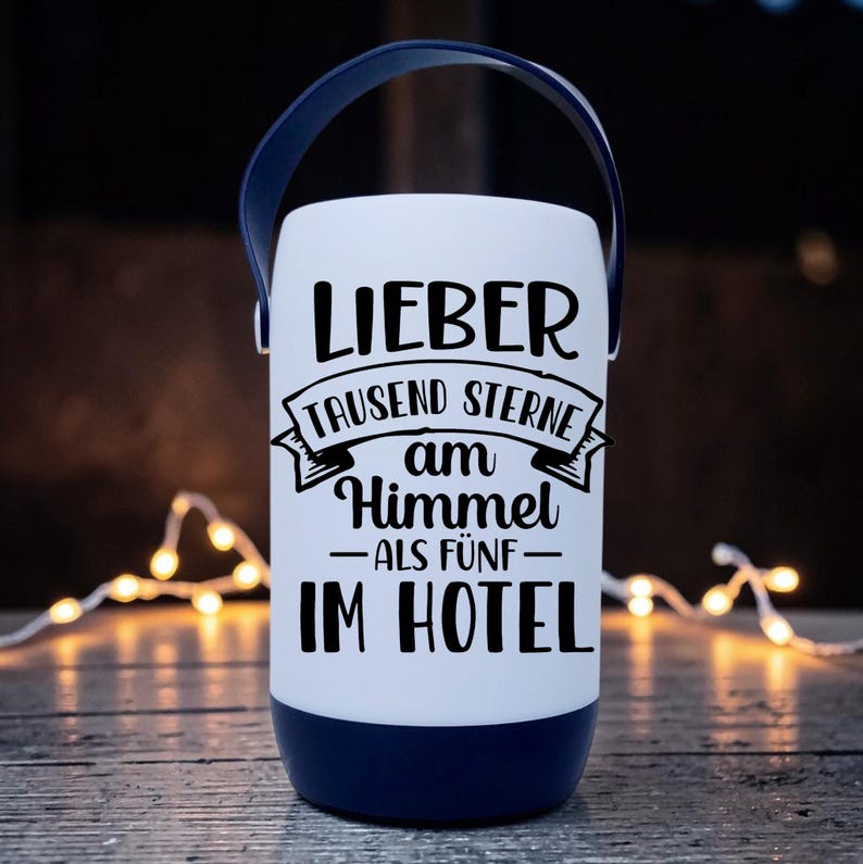 Peut inclure: Une lanterne blanche avec une poign&eacute;e et une base bleu marine. La lanterne porte un texte noir qui se lit "LIEBER TAUSEND STERNE AM HIMMEL ALS F&Uuml;NF IM HOTEL". La lanterne est pos&eacute;e sur une surface en bois avec des guirlandes lumineuses en arri&egrave;re-plan.