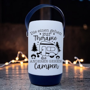 Peut inclure: Une lanterne de camping blanche et bleu marine avec une poign&eacute;e. La lanterne porte du texte et des graphiques noirs qui se lisent "Die einen gehen zur Therapie, die anderen gehen campen" avec une illustration de camping-car. La lanterne est cylindrique.