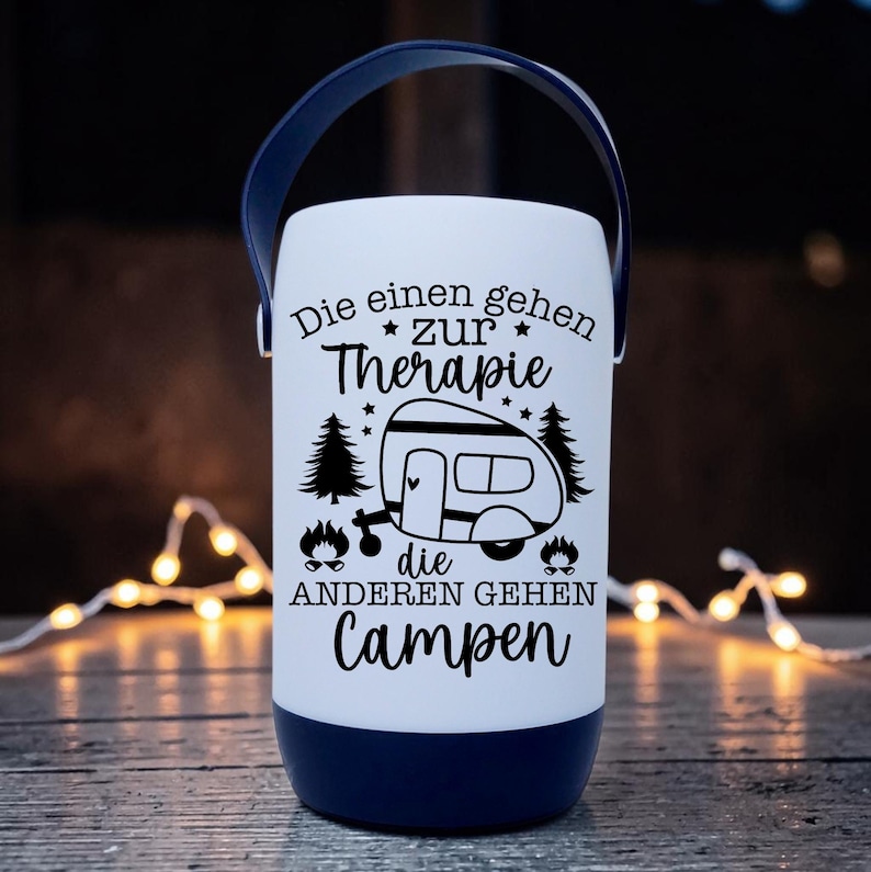 Peut inclure: Une lanterne de camping blanche et bleu marine avec une poign&eacute;e. La lanterne porte du texte et des graphiques noirs qui se lisent "Die einen gehen zur Therapie, die anderen gehen campen" avec une illustration de camping-car. La lanterne est cylindrique.