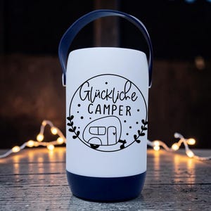 Peut inclure: Une lanterne de camping blanche et bleu marine avec une poign&eacute;e. La lanterne pr&eacute;sente le texte "Gl&uuml;ckliche Camper" dans un design circulaire noir avec une illustration de camping-car. La lanterne est cylindrique.