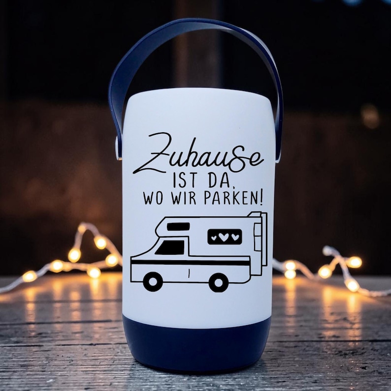Peut inclure: Une lanterne blanche et bleu marine avec une poign&eacute;e. La lanterne porte un dessin noir d'une caravane et le texte "Zuhause ist da, wo wir parken !" La lanterne est &eacute;clair&eacute;e.