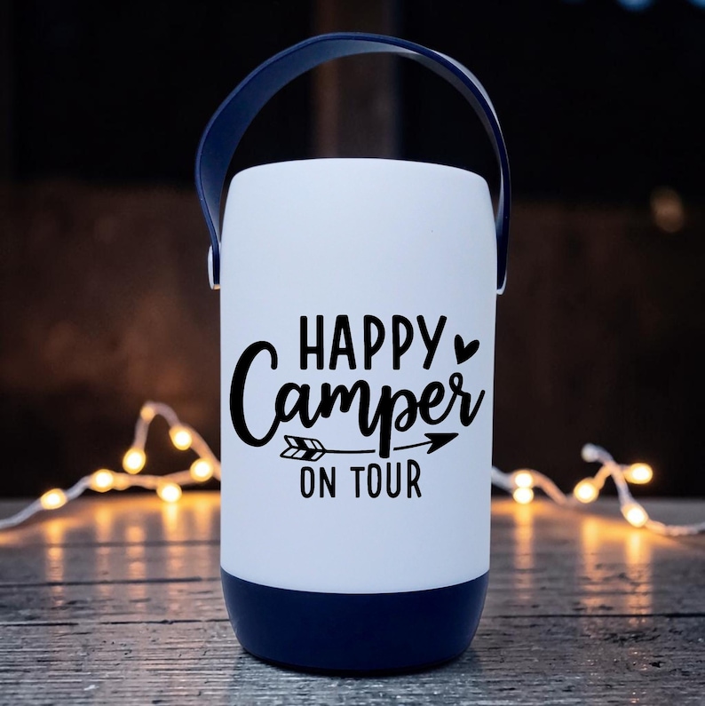 Peut inclure: Une lanterne de camping blanche et bleu marine avec une poign&eacute;e. La lanterne porte l'inscription "HAPPY Camper ON TOUR" en noir. La lanterne est cylindrique et poss&egrave;de une base et une poign&eacute;e bleu marine.