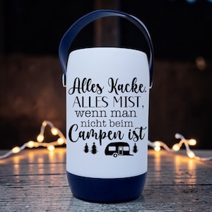 Peut inclure: Une lanterne de camping blanche et bleu marine avec une poign&eacute;e bleu marine. La lanterne porte un texte noir qui se lit "Alles Kacke, ALLES MIST, wenn man nicht beim Campen ist" avec un dessin de caravane. La lanterne est sur une surface en bois.