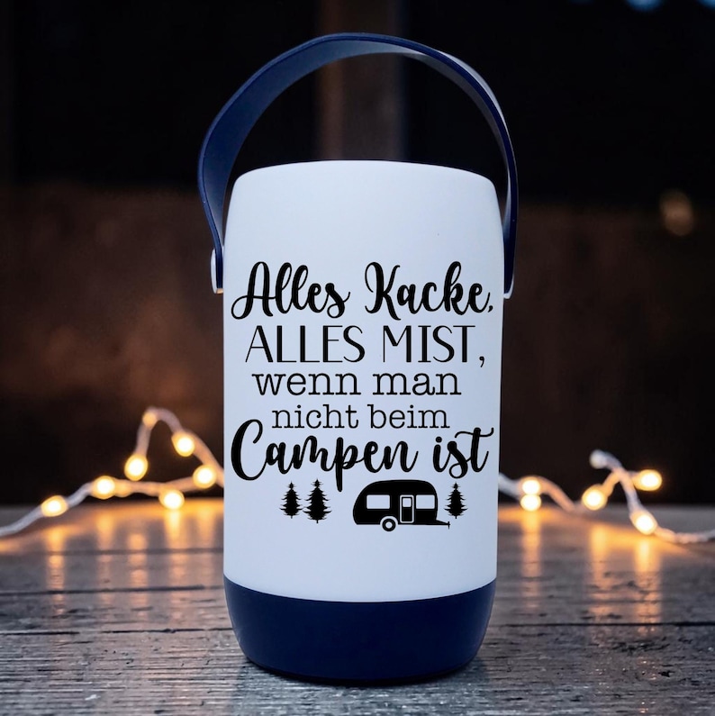 Peut inclure: Une lanterne de camping blanche et bleu marine avec une poign&eacute;e bleu marine. La lanterne porte un texte noir qui se lit "Alles Kacke, ALLES MIST, wenn man nicht beim Campen ist" avec un dessin de caravane. La lanterne est sur une surface en bois.