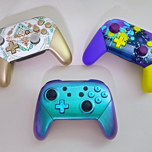 May include: Three custom game controllers. One is gold with a white and green design, another has a purple and yellow colour scheme with paint splatter, and the third is teal and purple.