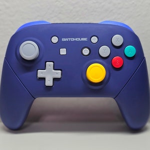 Peut inclure: Une manette Nintendo Switch bleue et grise avec un bouton jaune et le texte "SWITCHHOUSE" imprimé sur le devant.