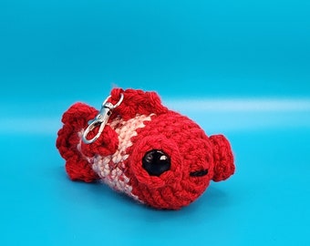 May include: A red and white crocheted betta fish keychain with black eyes. The keychain has a silver metal clasp.