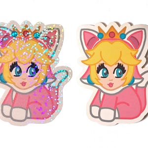 May include: Two stickers featuring a cartoon character wearing a pink dress, a crown, and cat ears. One sticker has a glittery finish, while the other is a solid colour.