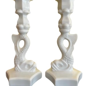 May include: A pair of white ceramic candlestick holders shaped like fish. The holders have a scalloped top and a fish tail base.