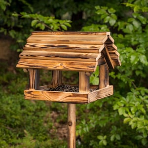 May include: A wooden bird feeder with a peaked roof and a square base, mounted on a wooden post. The bird feeder has a natural wood grain finish with dark brown accents. The background is blurred green foliage.
