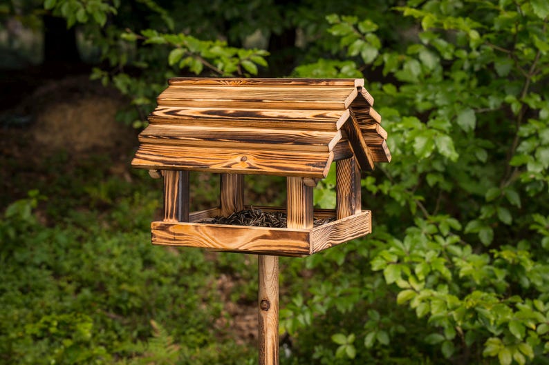 May include: A wooden bird feeder with a peaked roof and a square base, mounted on a wooden post. The bird feeder has a natural wood grain finish with dark brown accents. The background is blurred green foliage.