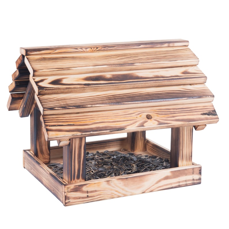 May include: A wooden bird feeder with a pitched roof, filled with sunflower seeds. The feeder has a natural wood grain finish with darker, burnt-looking accents. The design is reminiscent of a small house.