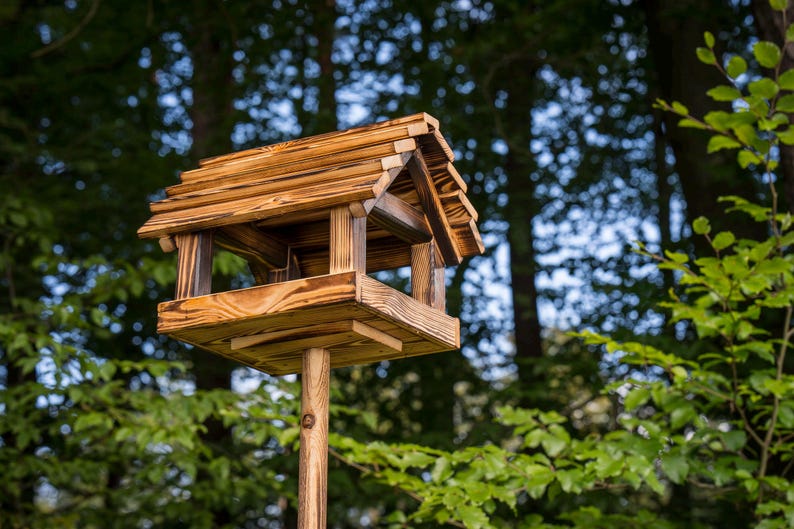 May include: A wooden birdhouse with a pitched roof and a square base, mounted on a wooden pole. The birdhouse is made of light brown wood and has a natural finish. The background features lush green foliage and trees.