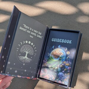 May include: Open tarot card box with a guidebook. The box is dark with a tree of life design and a quote by Rumi. The guidebook cover features a woman in a green dress, a full moon, and colorful flowers. The word "GUIDEBOOK" is printed on the cover.