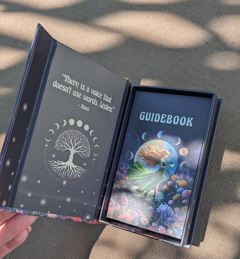 May include: Open tarot card box with a guidebook. The box is dark with a tree of life design and a quote by Rumi. The guidebook cover features a woman in a green dress, a full moon, and colorful flowers. The word "GUIDEBOOK" is printed on the cover.