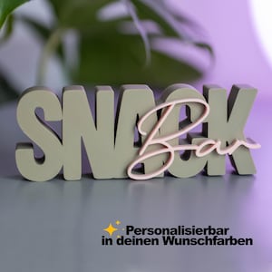 Könnte beinhalten: Ein dreidimensionales Schild mit dem Wort SNACK in großen, salbeigrünen Blockbuchstaben, wobei das Wort Bar in einer hellrosa Schreibschrift darüber liegt. Darunter steht der Text Personalisierbar in deinen Wunschfarben.