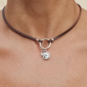 Puede incluir: Un collar gargantilla marrón con un anillo plateado y un pequeño colgante con una piedra transparente de talla cuadrada. El collar se lleva alrededor del cuello.