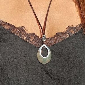 Peut inclure: Un collier avec un pendentif argenté en forme de goutte d'eau. Le pendentif est suspendu à un cordon en cuir marron. Le collier est porté sur un haut noir.