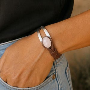 Op de afbeelding: Een armband met een gladde, ovaalvormige lichtroze steen in een zilverkleurige metalen zetting, bevestigd aan een donkerbruine leren band die om de pols wikkelt, afgewerkt met een eenvoudig knoopdetail bij de centrale steen.