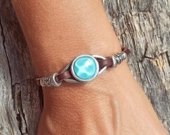 May include: A bracelet featuring a dark brown leather cord accented with silver-toned metal components and a central circular turquoise-colored cabochon. The design includes decorative silver-toned beads on each side of the central stone setting.