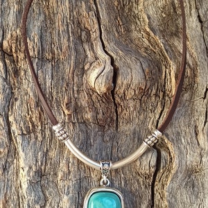 May include: A brown leather necklace with a silver pendant featuring a turquoise-colored stone. The pendant has a decorative silver band around the stone and a small silver bead hanging from the bottom.