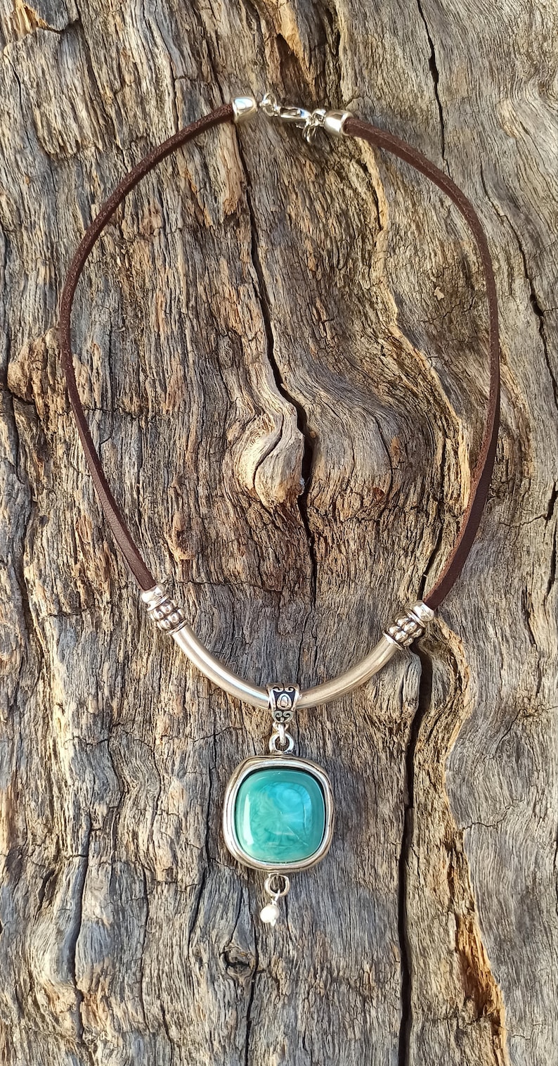 May include: A brown leather necklace with a silver pendant featuring a turquoise-colored stone. The pendant has a decorative silver band around the stone and a small silver bead hanging from the bottom.