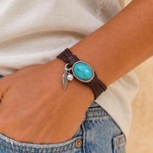 Könnte beinhalten: Ein mehrsträngiges dunkelbraunes Lederarmband mit einem zentralen ovalen Türkisstein in einer silberfarbenen Fassung. Das Armband enthält zwei hängende Anhänger: eine kleine silberfarbene Blattform und eine einzelne weiße Perle.